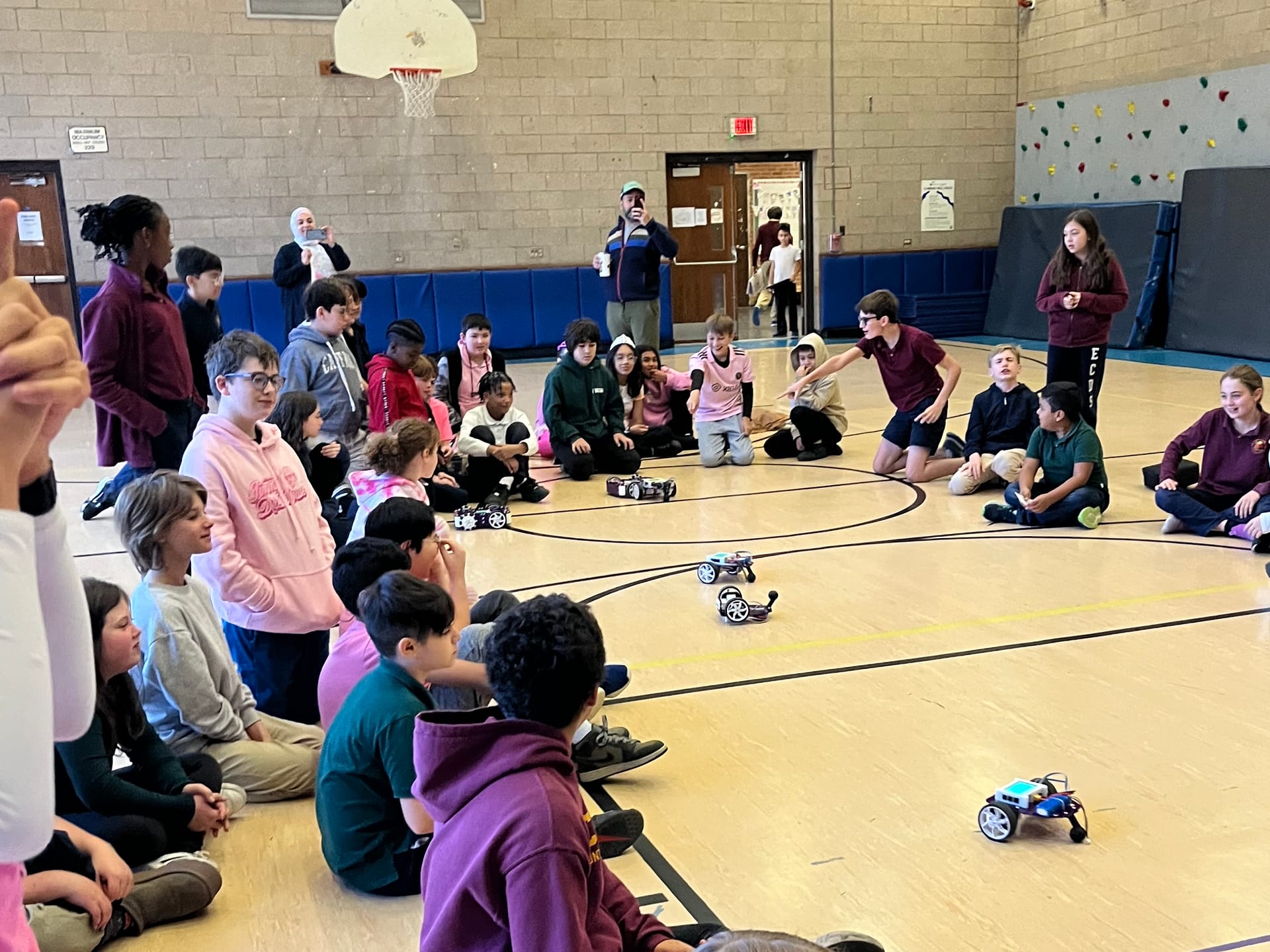 Students sit and stand around the edge of a gym floor and watch five robot kits drive between them