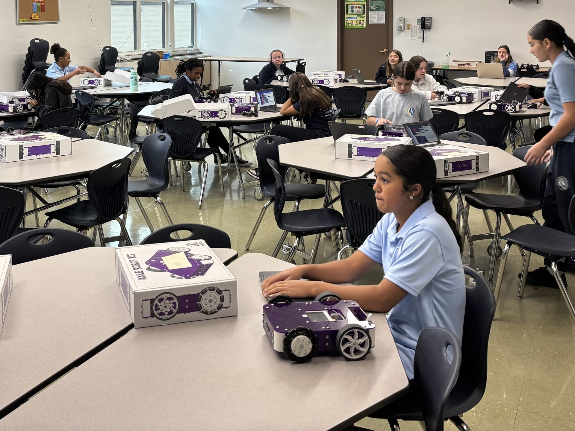 Middle school students at classroom tables open Axle Robot Kit boxes with a built robot in the foreground