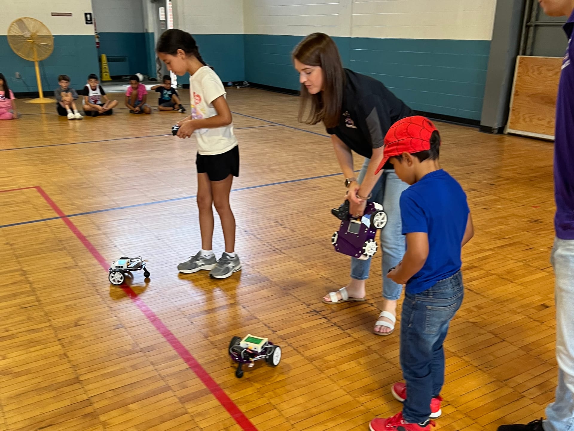 Two young students drive robots across a gym floor while a True Robotics instructor holds an Axle robot and leans in to help one of them