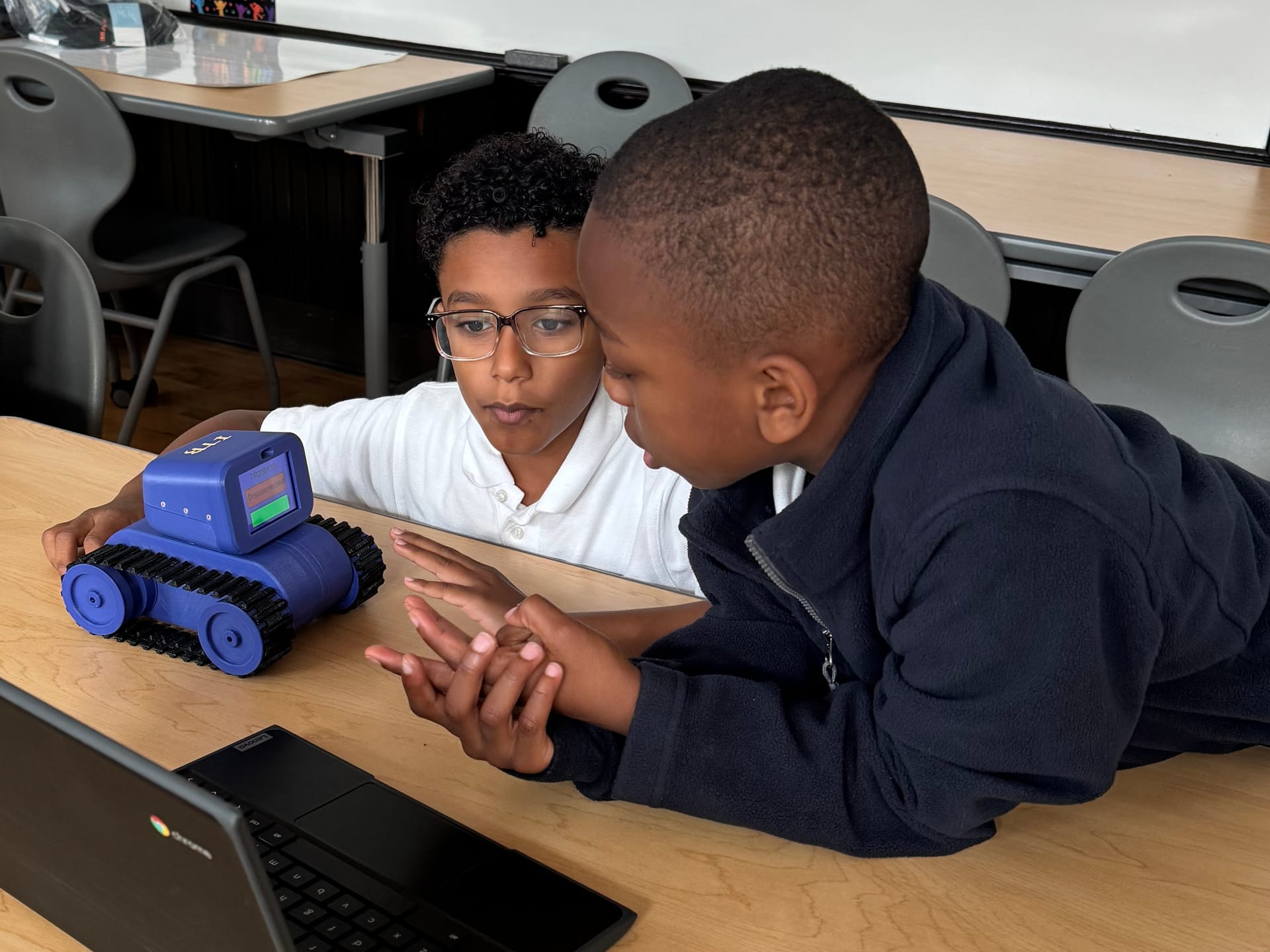 Two young students lean across a classroom table and talk it over beside a Treads Robot and their Chromebook