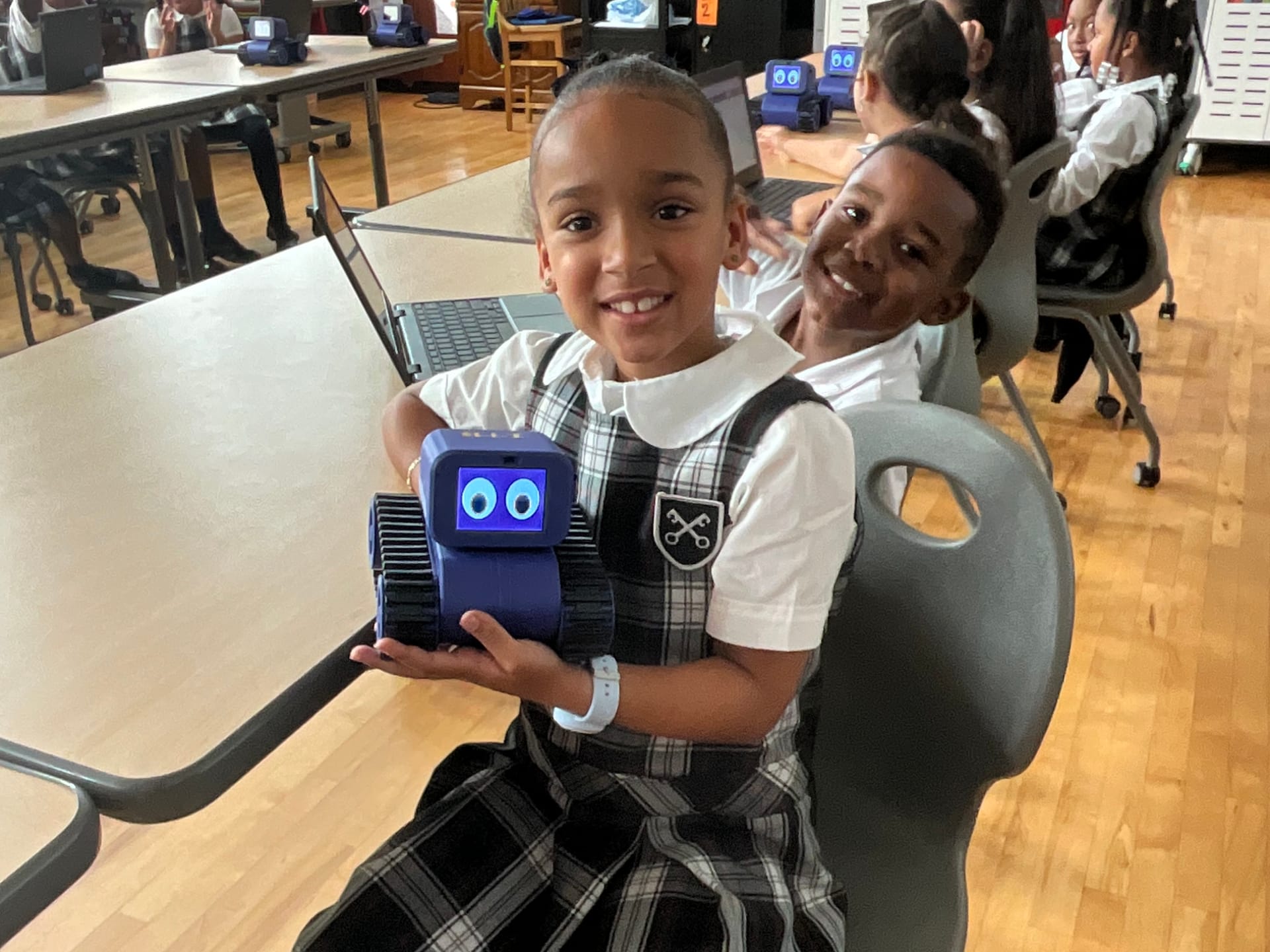 A young student smiles as she holds up a Treads Robot with cartoon eyes on its screen while her classmates work at tables behind her