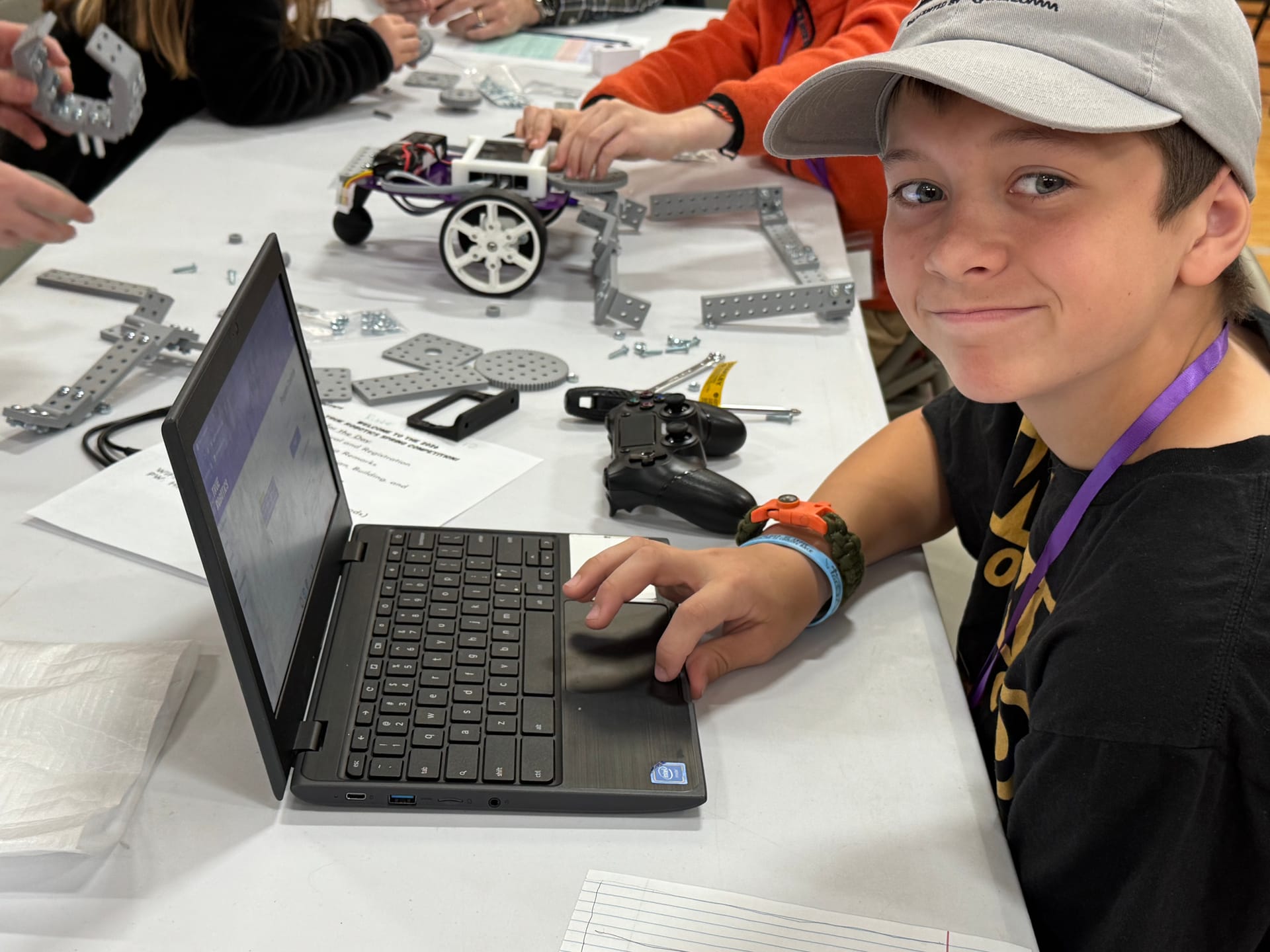 A student smiles at a Chromebook while programming a Pilot robot fitted with FlexForge beams at a competition table