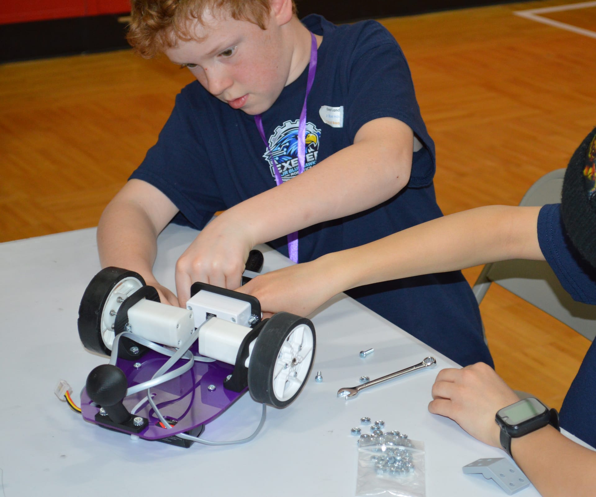 A student fastens parts onto a purple Pilot robot base with a classmate, a wrench, and a bag of nuts beside them on the table