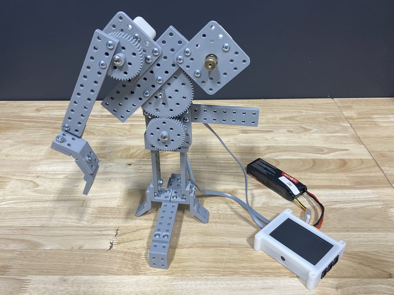 A gray FlexForge prosthetic arm with gears and a gripper hand stands on a wooden table, wired to a white robot controller and a battery