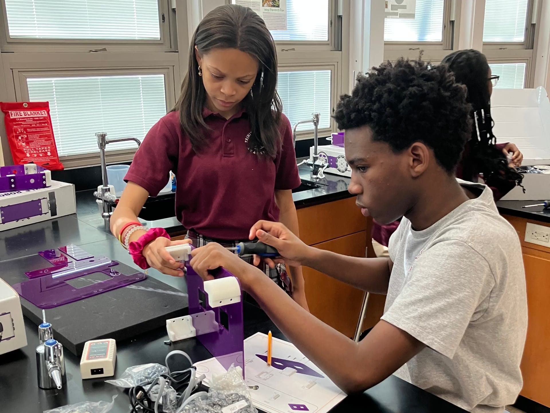 Two students at a science lab bench build a purple robot kit together, one turning a screwdriver while the other holds a part beside the printed guide