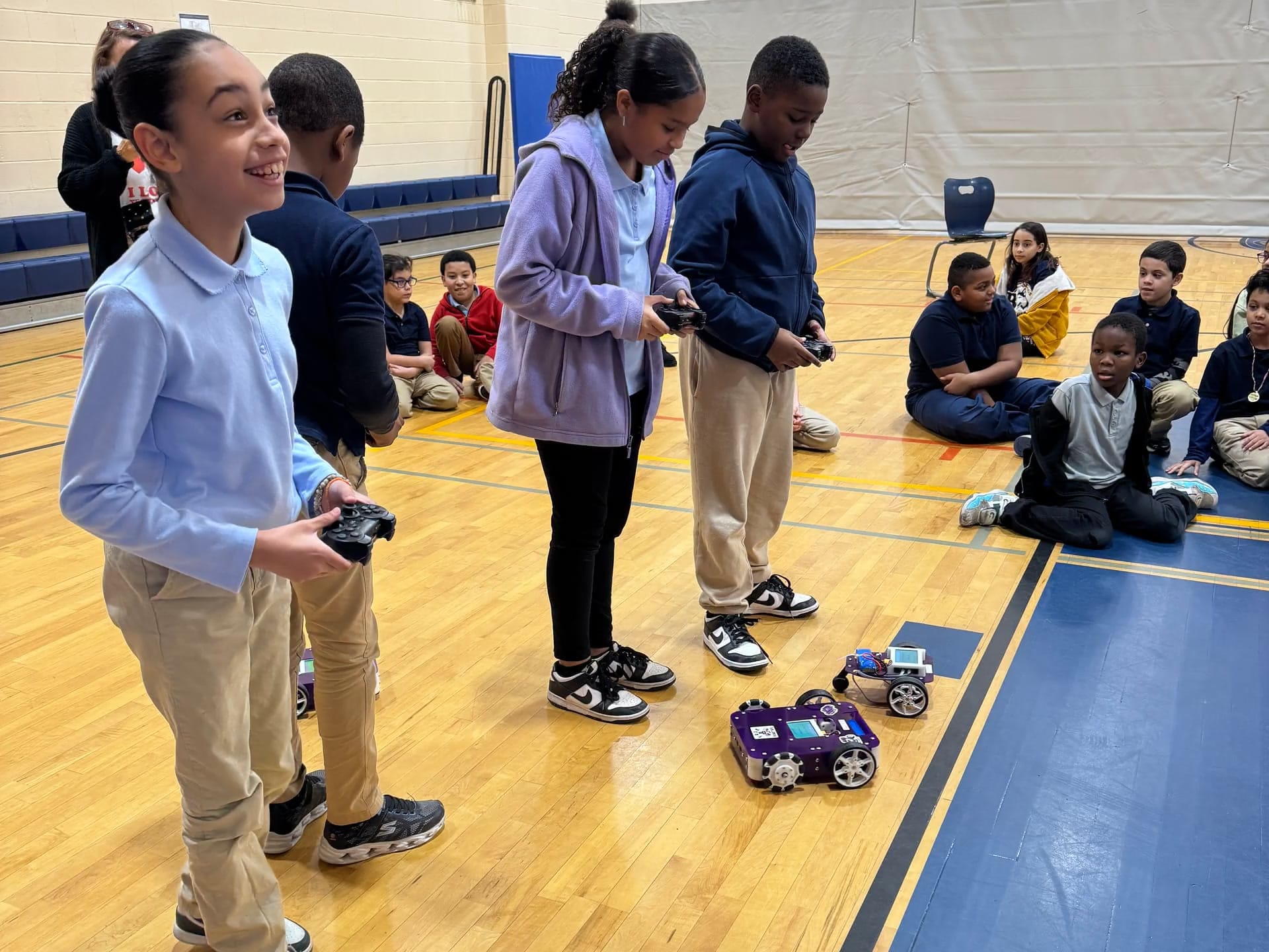 Three elementary students smile as they steer Axle and Pilot robots with game controllers while classmates watch from the gym floor