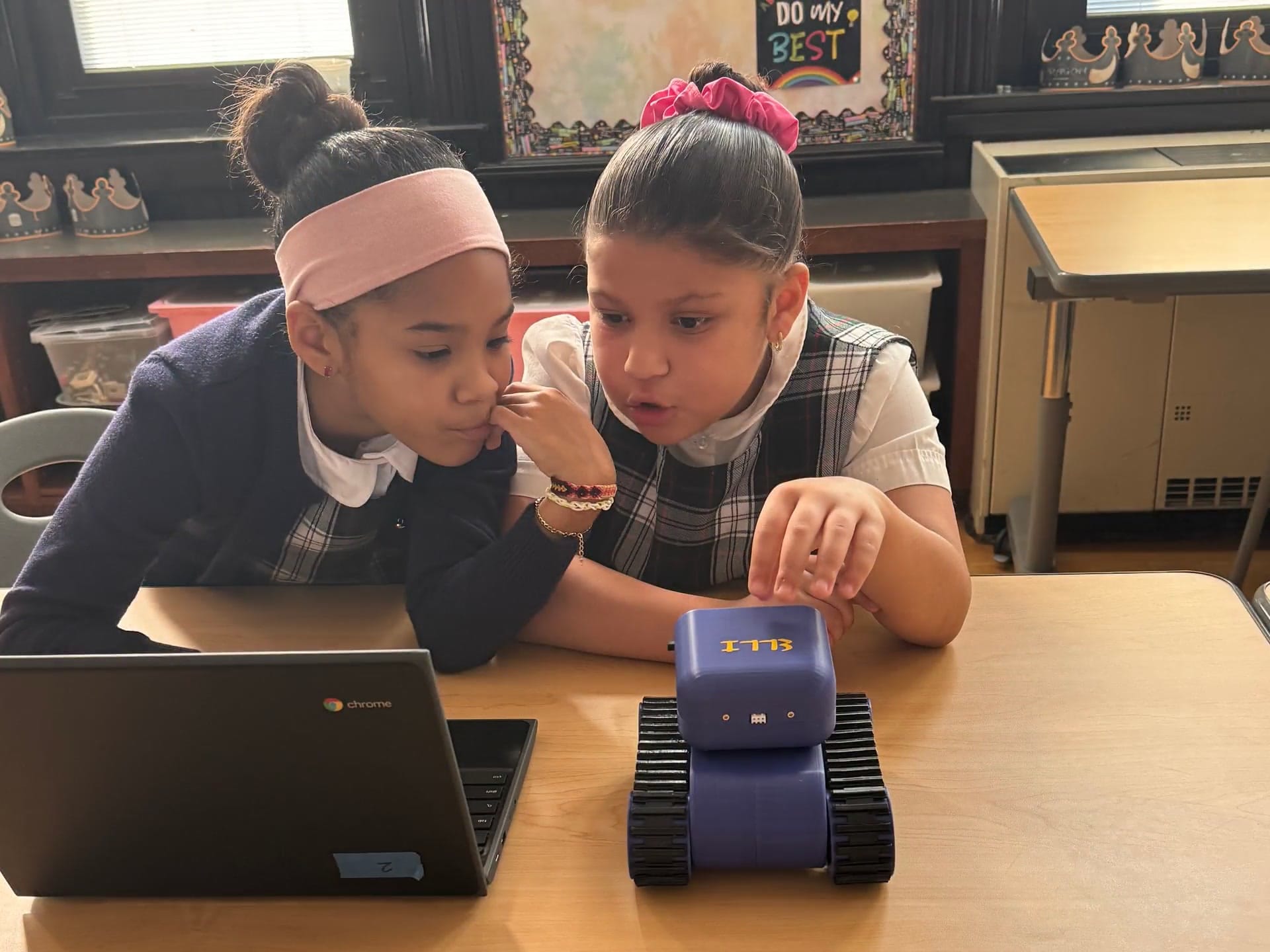 Two young students lean in over a classroom table as one reaches for a Treads Robot beside their Chromebook